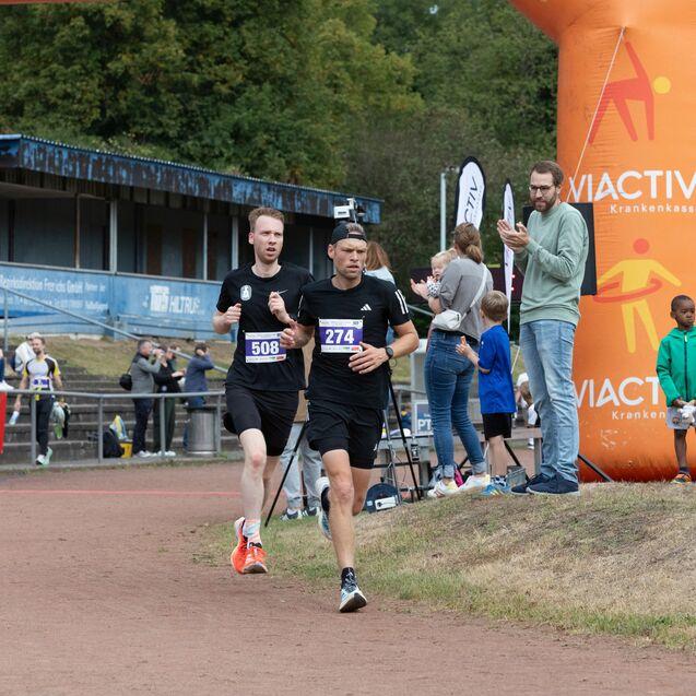 Zwei L&auml;ufer in Sportbekleidung &uuml;berqueren die Ziellinie eines laufenden Events, w&auml;hrend Zuschauer applaudieren.