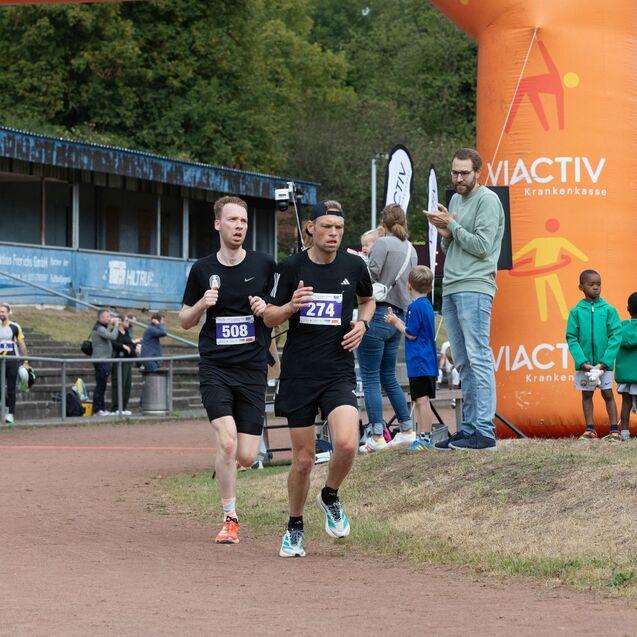 Zwei L&auml;ufer mit Startnummern 508 und 274 passieren den Zielbogen bei einem Wettkampf, umgeben von Zuschauern.