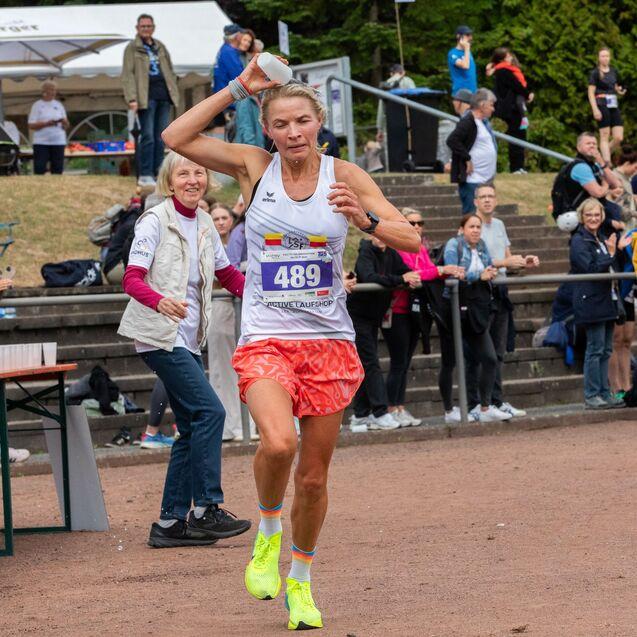 L&auml;uferin mit Startnummer 489 beugt sich vor, w&auml;hrend sie ein Wasserflasche h&auml;lt, umgeben von jubelnden Zuschauern.