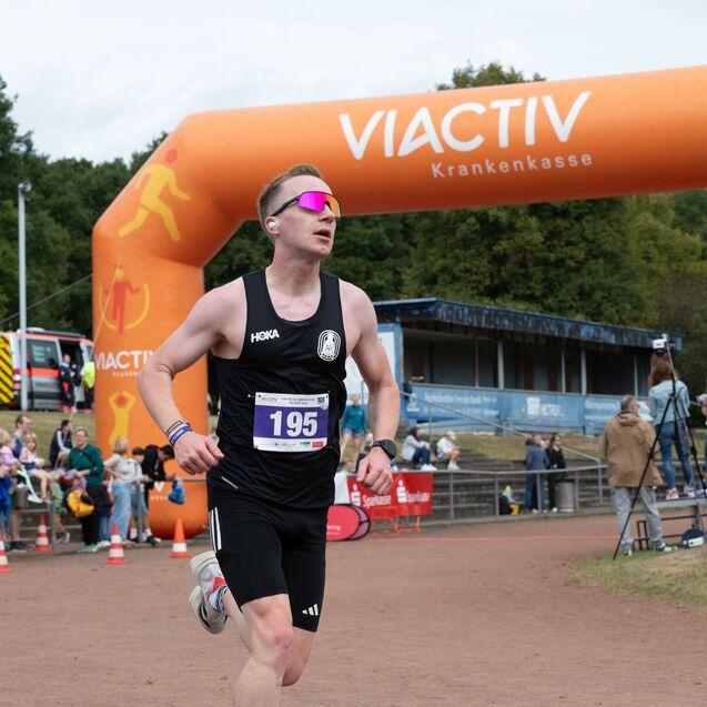 L&auml;ufer mit Startnummer 195 sprintet unter einem orangefarbenen Zielbogen bei einem Wettkampf, Zuschauer im Hintergrund.