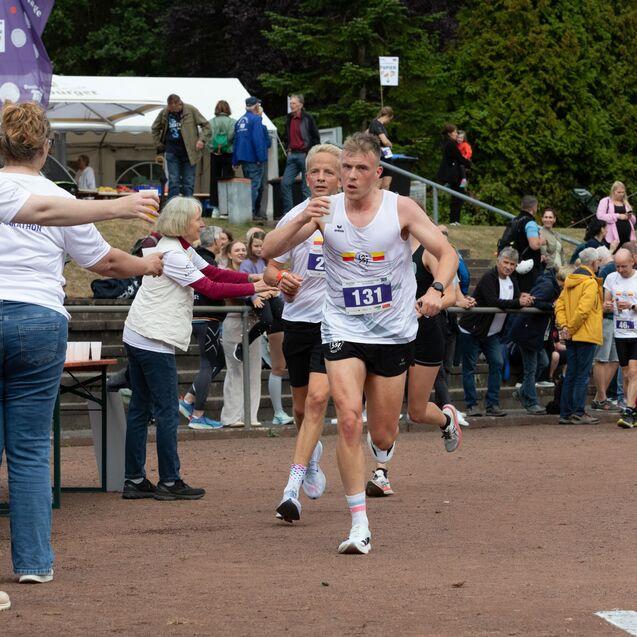 Zwei L&auml;ufer &uuml;bergeben w&auml;hrend eines Halbmarathons einen Staffelwechsel an einem Verpflegungspunkt, umgeben von Zuschauern.