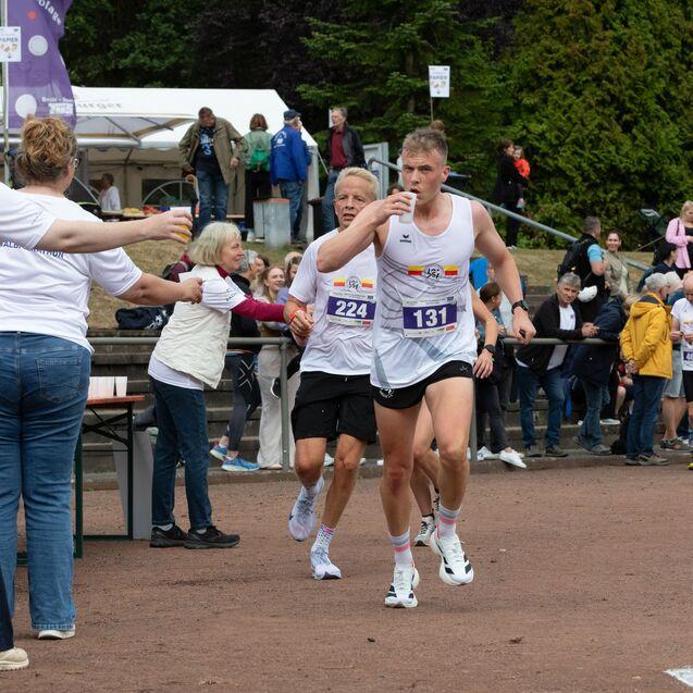 L&auml;ufer &uuml;bergibt den Staffelstab w&auml;hrend des Halbmarathons an einen Teamkollegen, umgeben von Zuschauern und Helfern.