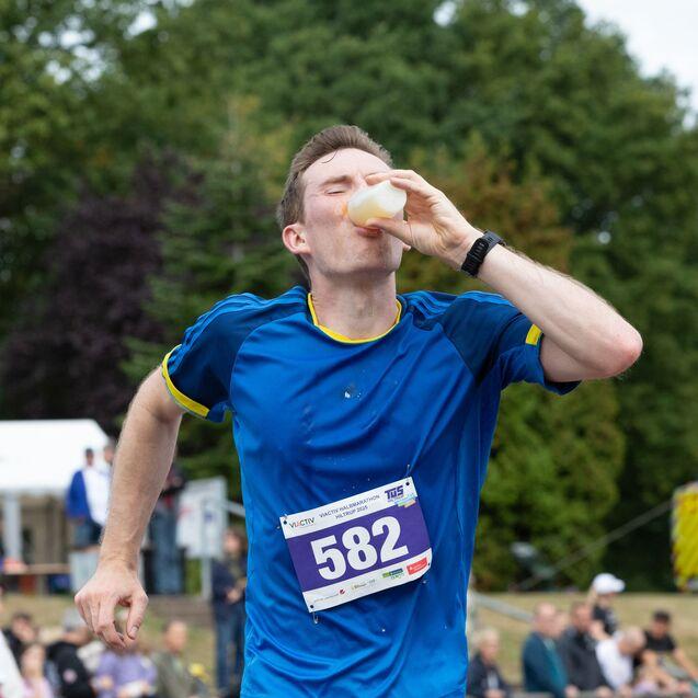 L&auml;ufer in blauer Sportkleidung trinkt aus einer Becherh&auml;lfte w&auml;hrend eines Wettkampfs im Freien.