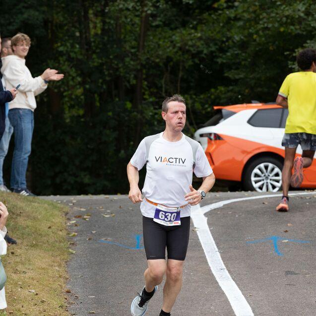 Teilnehmer eines Laufs l&auml;uft auf einer Stra&szlig;e, w&auml;hrend Zuschauer applaudieren. Im Hintergrund ein orangefarbenes Auto.