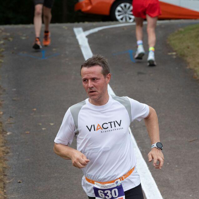 L&auml;ufer in wei&szlig;em Shirt mit &bdquo;VIACTIV&ldquo; und Nummer 630, unterwegs auf einer Laufstrecke mit grauem Asphalt.