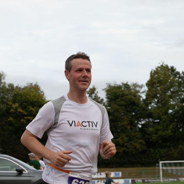 Mann in einem wei&szlig;en T-Shirt beim Laufen auf einem Sportgel&auml;nde, im Hintergrund B&auml;ume und Zuschauer.