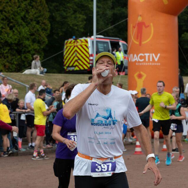 L&auml;ufer in wei&szlig;em Shirt trinkt aus einer Flasche w&auml;hrend eines Wettkampfs, im Hintergrund Publikum und Anfeuerungen.
