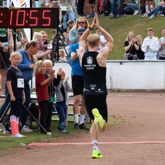 L&auml;ufer mit erhobenen H&auml;nden &uuml;berquert die Ziellinie, w&auml;hrend Kinder und Zuschauer jubeln; Zeit 1:10:55 auf der Anzeige.