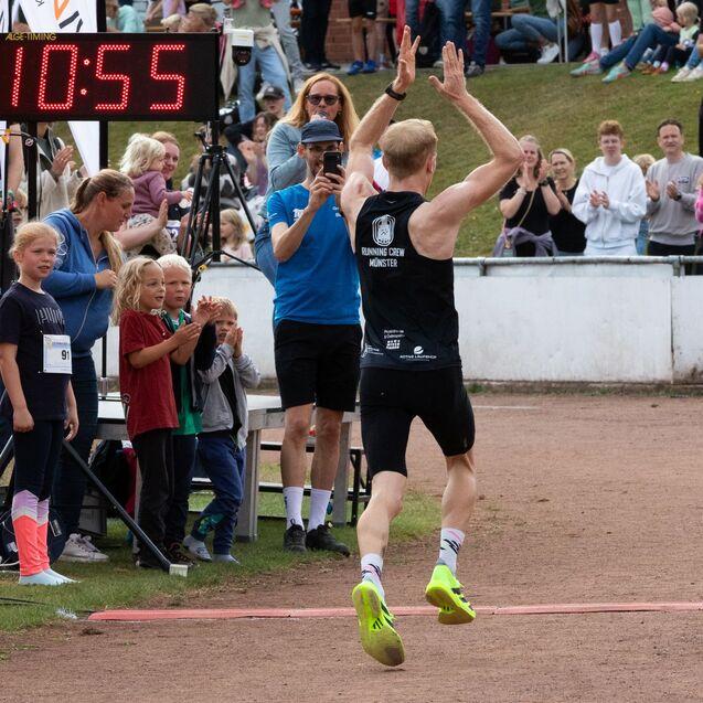 L&auml;ufer mit hochgestreckten Armen jubelt nach dem Ziel in einer Laufveranstaltung, Zuschauer applaudieren im Hintergrund.