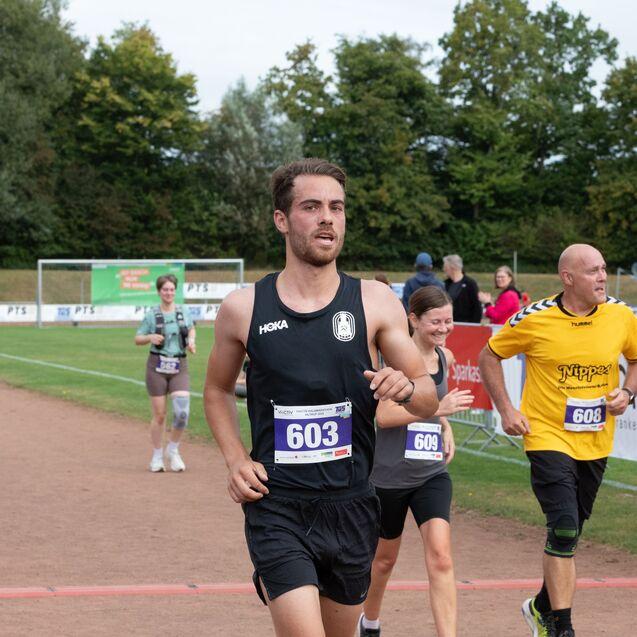 Ein L&auml;ufer in schwarzem Shirt n&auml;hert sich der Ziellinie bei einem Wettkampf, umgeben von weiteren Teilnehmern.