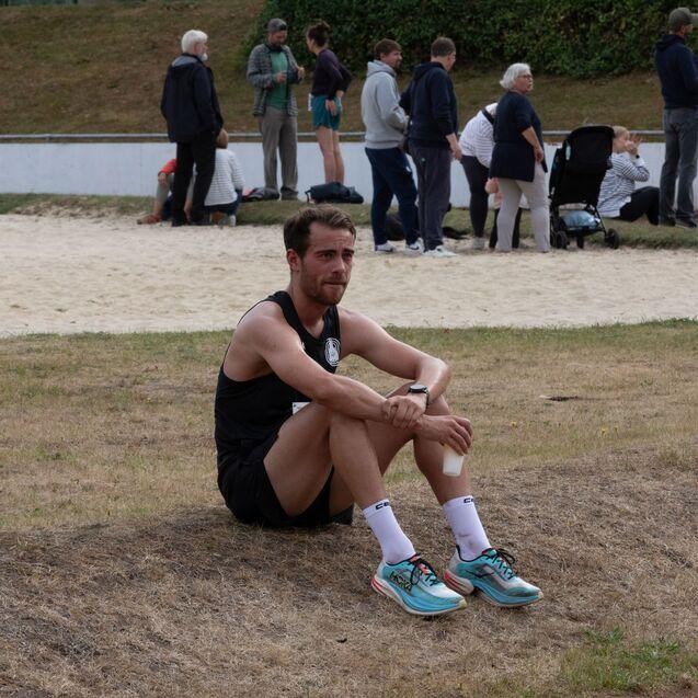 Ein junger Sportler in Shorts sitzt auf dem Boden neben einer Sandfl&auml;che, umgeben von Zuschauergruppen.