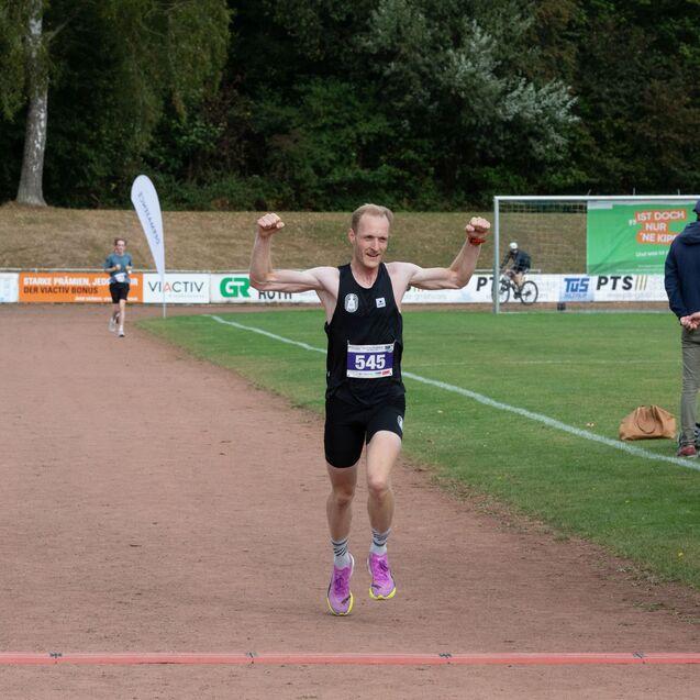 L&auml;ufer mit der Nummer 545 jubelt beim &Uuml;berqueren der Ziellinie auf einer Sportanlage.