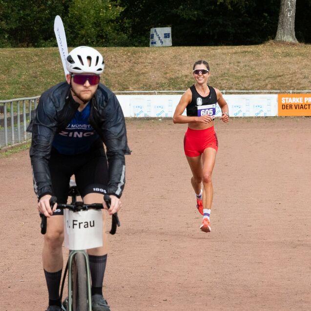 Eine L&auml;uferin in sportlicher Kleidung l&auml;uft auf einer Laufbahn, w&auml;hrend ein Radfahrer neben ihr f&auml;hrt.
