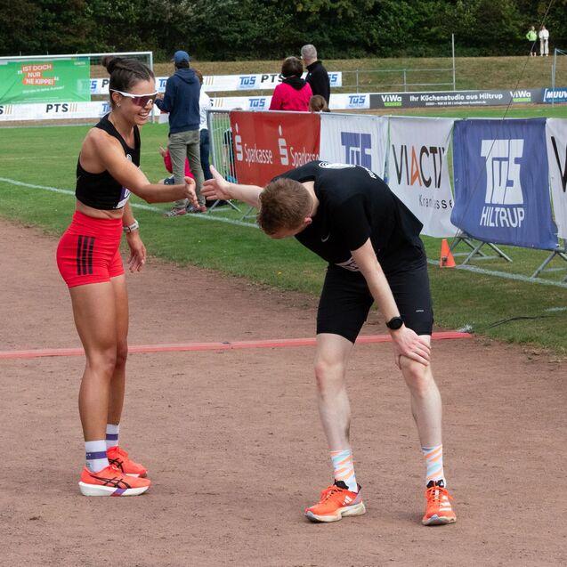 Sportlerin gratuliert ersch&ouml;pftem L&auml;ufer nach Wettkampf auf Laufbahn, im Hintergrund Zuschauer und Werbebanner.