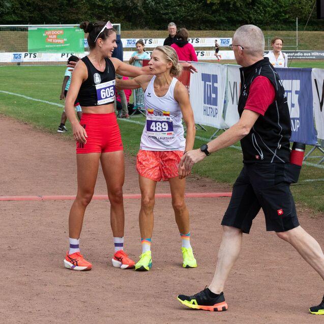 Zwei Athletinnen in Sportbekleidung umarmen sich nach einem Wettkampf, ein Funktion&auml;r beobachtet die Szene.