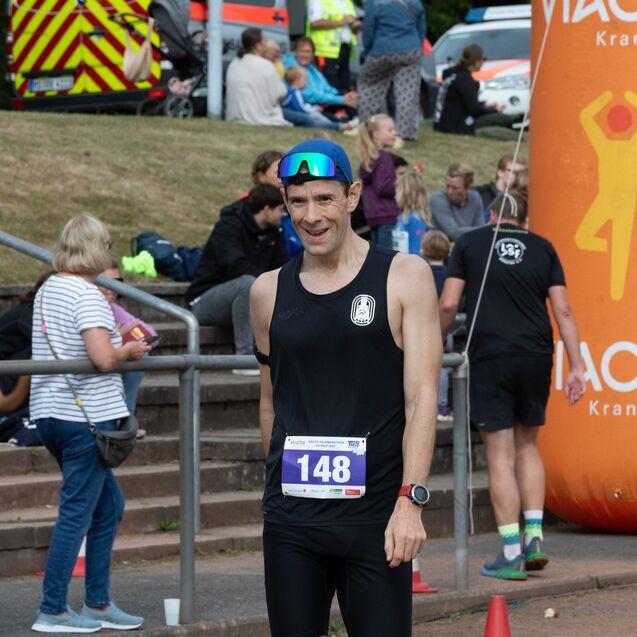 L&auml;ufer in Sportkleidung mit Startnummer 148 steht l&auml;chelnd vor einem Orangenfu&szlig;ball-Torbogen und Zuschauern im Hintergrund.