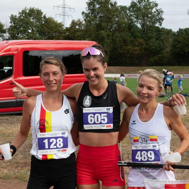 Drei Frauen in Sportbekleidung posieren nach einem Laufereignis, im Hintergrund ein roter Transporter und Zuschauer.