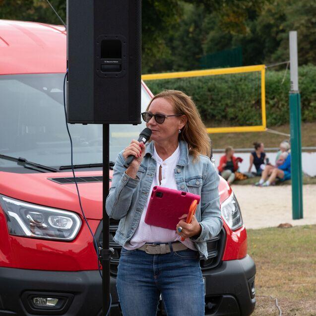 Frau mit Mikrofon und Tablet spricht vor einer Gruppe, im Hintergrund ein roter Transporter und ein Volleyballfeld.