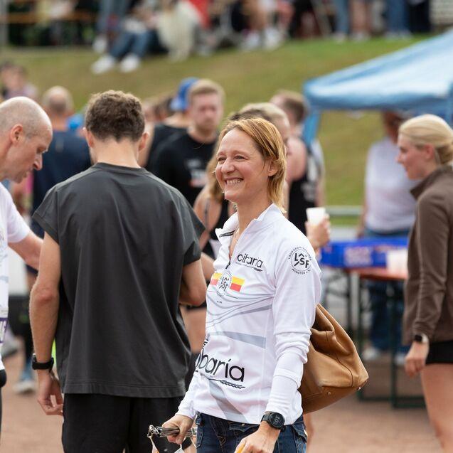 L&auml;chelnde Frau mit kurz geschnittenem Haar in Sportbekleidung steht l&auml;ssig inmitten von Teilnehmern einer Sportveranstaltung.
