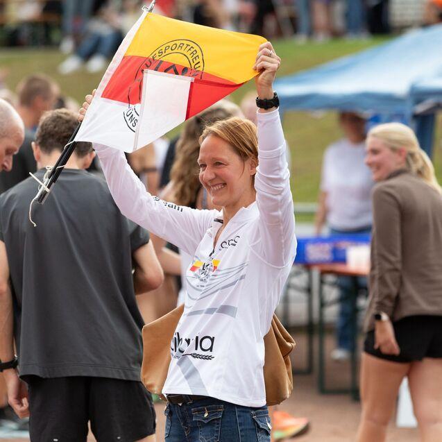 Frau hebt ein Flagge mit Vereinswappen &uuml;ber einer Menge bei einem Sportevent. Im Hintergrund sind Teilnehmer und Zelte zu sehen.