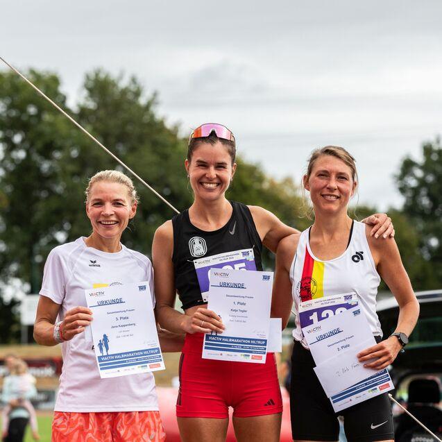 Drei Frauen stehen l&auml;chelnd auf einem Podium mit Urkunden nach einem Laufwettbewerb, im Hintergrund B&auml;ume und eine Flagge.