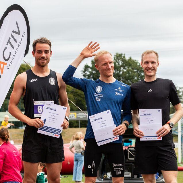 Drei L&auml;ufer stehen auf dem Podium, halten Urkunden f&uuml;r die Pl&auml;tze 1 bis 3 fest und l&auml;cheln in die Kamera.