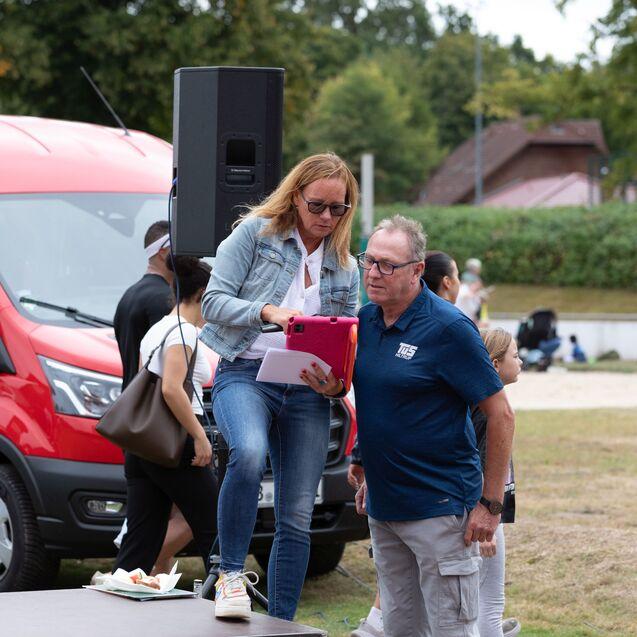Eine Frau steht auf einer Plattform und bedient ein Tablet, w&auml;hrend ein Mann danebenstehend schaut. Hintergrund mit Menschen und rotem Lieferwagen.
