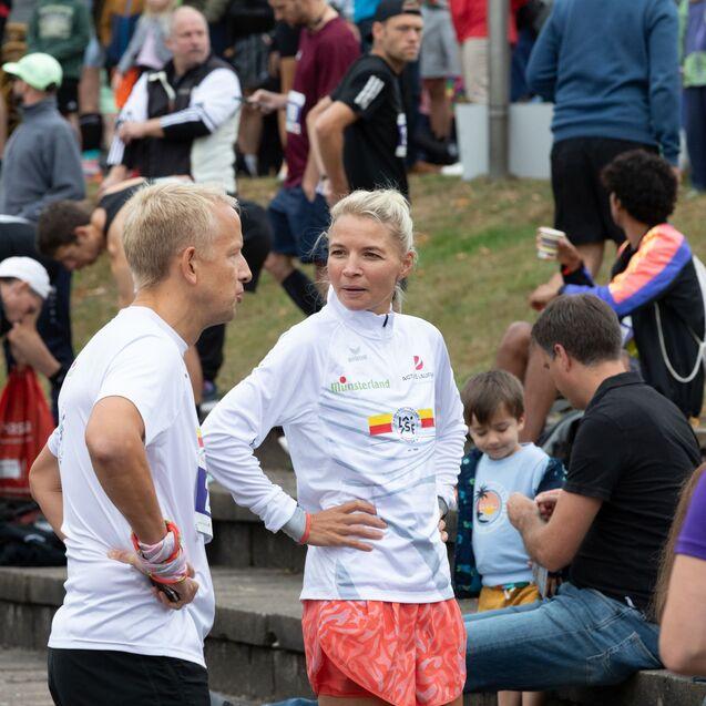 Zwei L&auml;ufer in Sportswear unterhalten sich, w&auml;hrend im Hintergrund weitere Teilnehmer auf den Wettbewerb warten.