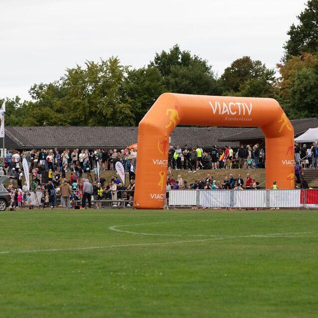 Gruppe von Menschen versammelt sich unter einem gro&szlig;en orangen Zielbogen auf einem Sportplatz im Freien.