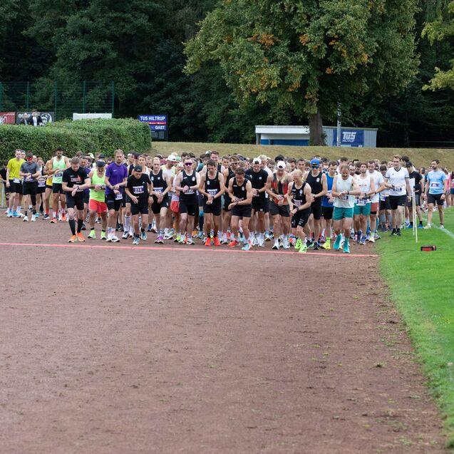 Gruppierung von L&auml;ufern an der Startlinie eines Wettlaufs auf einer Sportanlage, umgeben von Zuschauern und B&auml;umen.