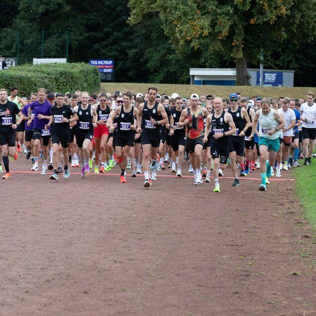 Gruppenauslauf von L&auml;ufern in schwarzer Sportbekleidung auf einer Laufbahn bei einem Wettkampf. Zuschauer sind im Hintergrund.