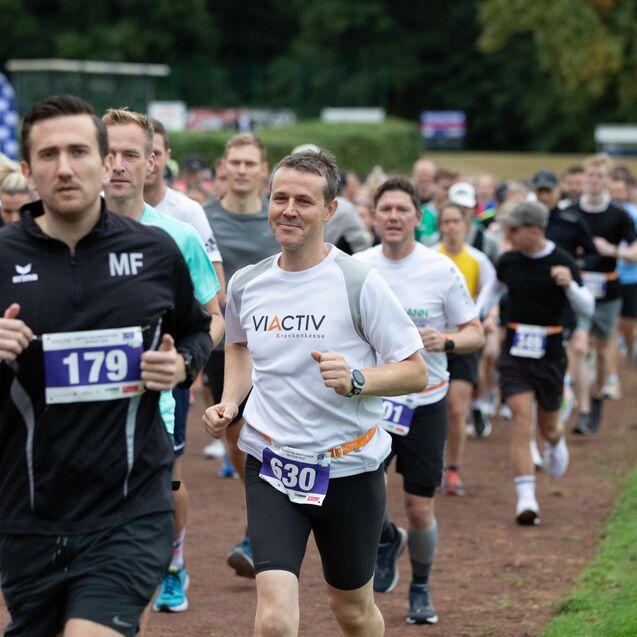 Gruppierung von Laufteilnehmern auf einer Rennstrecke, verschiedene Laufkleidungen und Startnummern sichtbar.