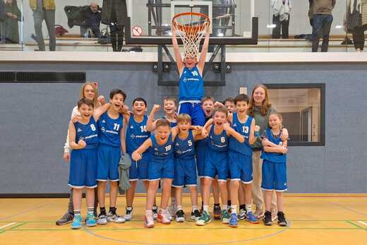 Jugend-Basketballmannschaft in blauen Trikots posiert fröhlich vor einem Korb in der Sporthalle.