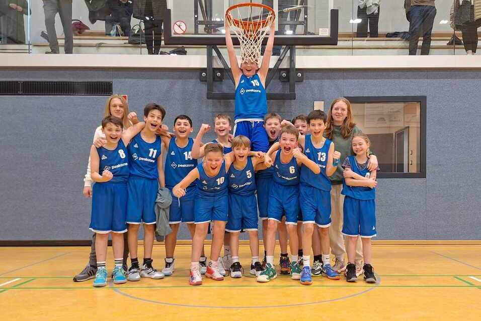 Jugend-Basketballmannschaft in blauen Trikots posiert fröhlich vor einem Korb in der Sporthalle.
