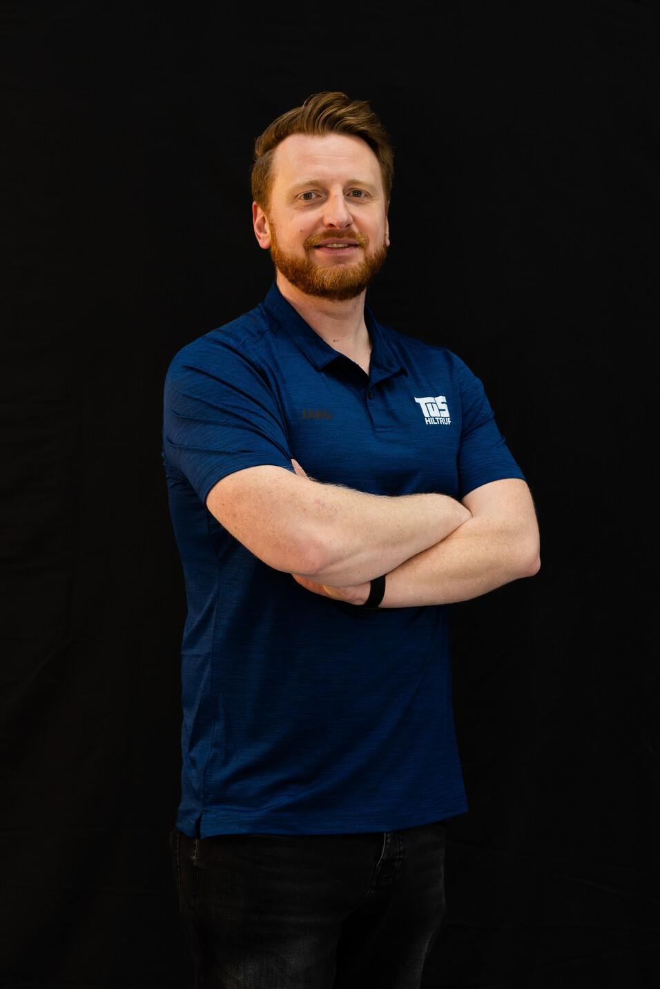 Mann in blauer Sportbekleidung mit verschränkten Armen vor schwarzem Hintergrund, lächelt leicht in die Kamera.