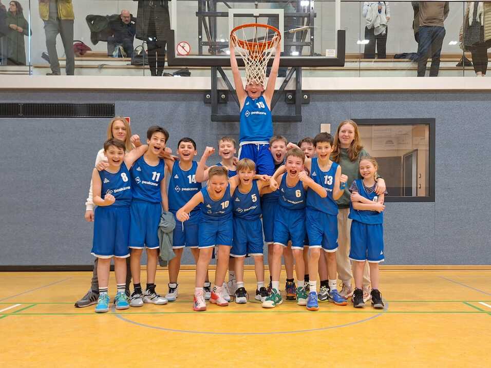 Jugend-Basketballmannschaft in blauen Trikots posiert fröhlich vor einem Korb in der Sporthalle.