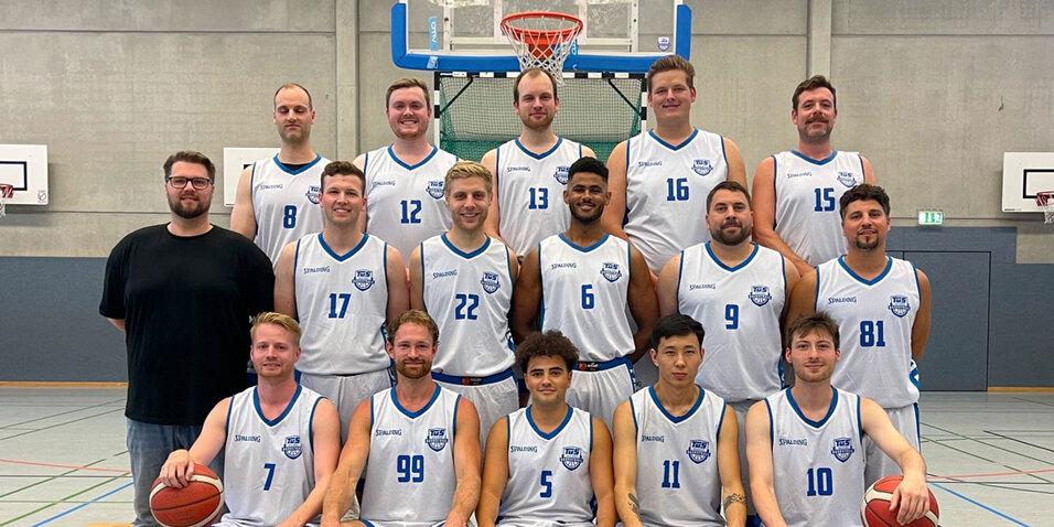Gruppenfoto einer Basketballmannschaft in Sportkleidung, stehend und sitzend in einer Sporthalle mit Basketballkorb.