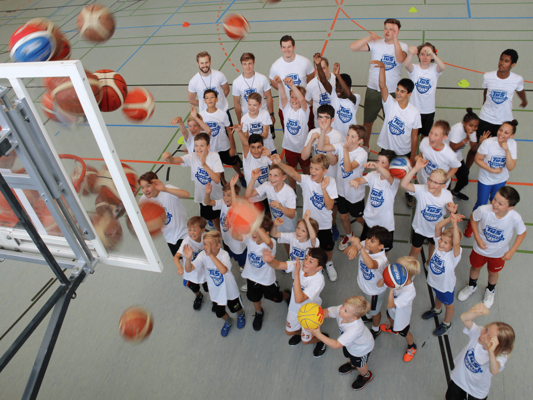 Gruppe von Kindern in wei&szlig;en T-Shirts, die auf einen Basketballkorb zielen, w&auml;hrend B&auml;lle in der Luft fliegen.