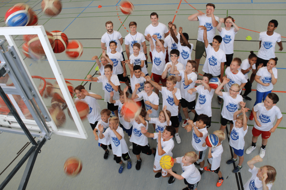 Gruppe von Kindern in weißen T-Shirts, die auf einen Basketballkorb zielen, während Bälle in der Luft fliegen.