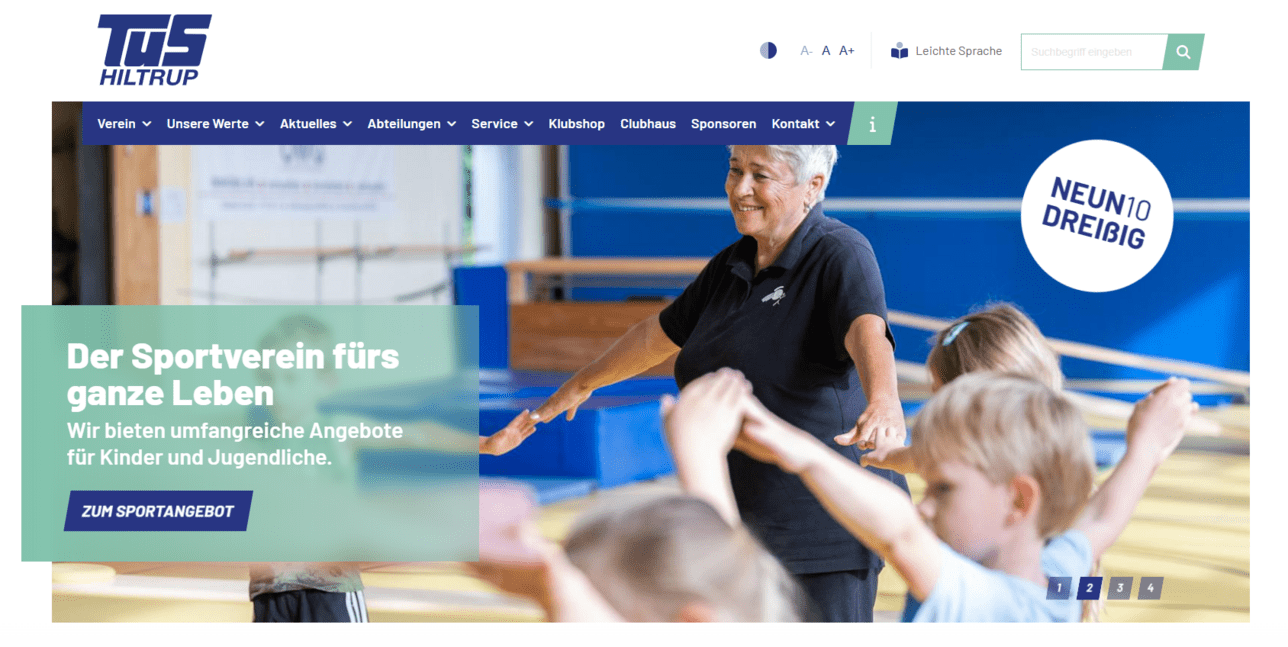 Frau in schwarzem T-Shirt demonstriert Übungen für Kinder in einem Sportverein. Hintergründe mit Turngeräten sichtbar.