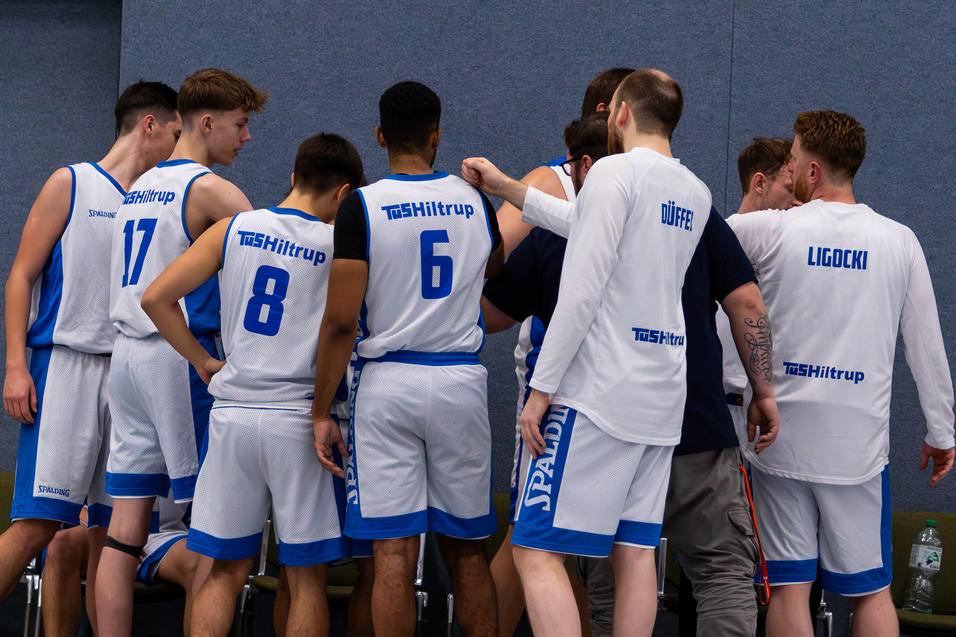 Mehrere Basketballspieler stehen in einem Huddle und besprechen strategie, w&auml;hrend auf der Bank im Hintergrund Flaschen stehen.