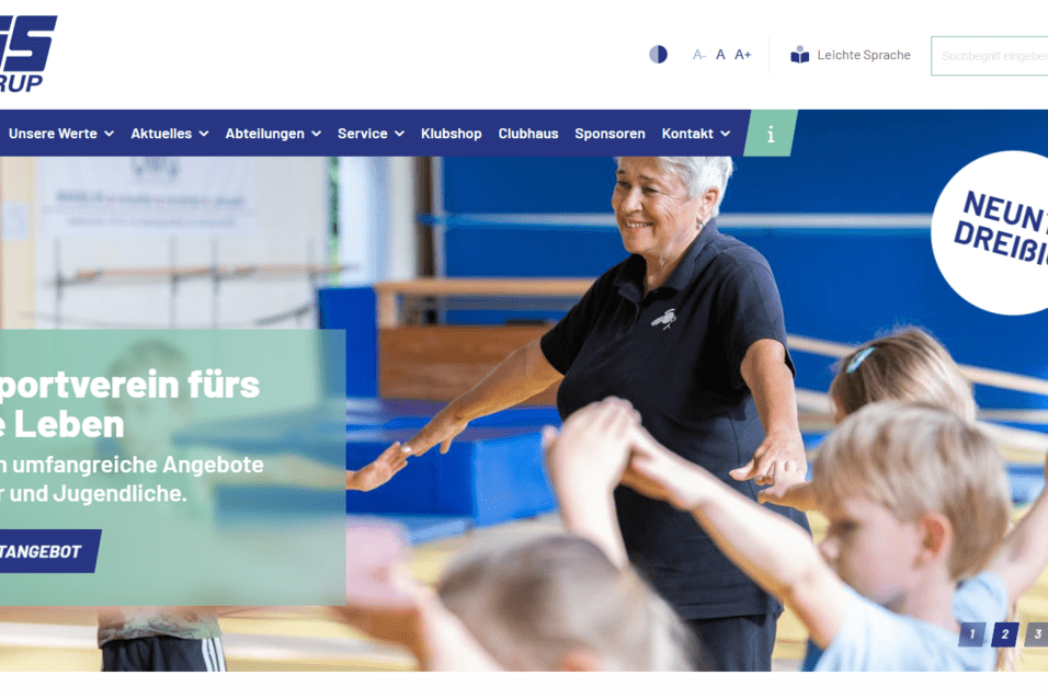 Frau in schwarzem T-Shirt demonstriert Übungen für Kinder in einem Sportverein. Hintergründe mit Turngeräten sichtbar.