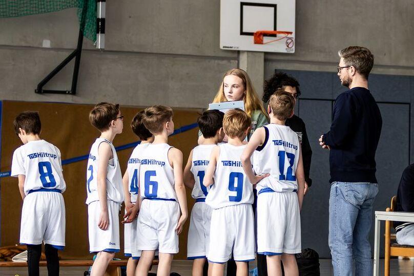 Gruppe junger Basketballspieler in weißer Sportkleidung versammelt sich um einen Coach in einer Sporthalle.