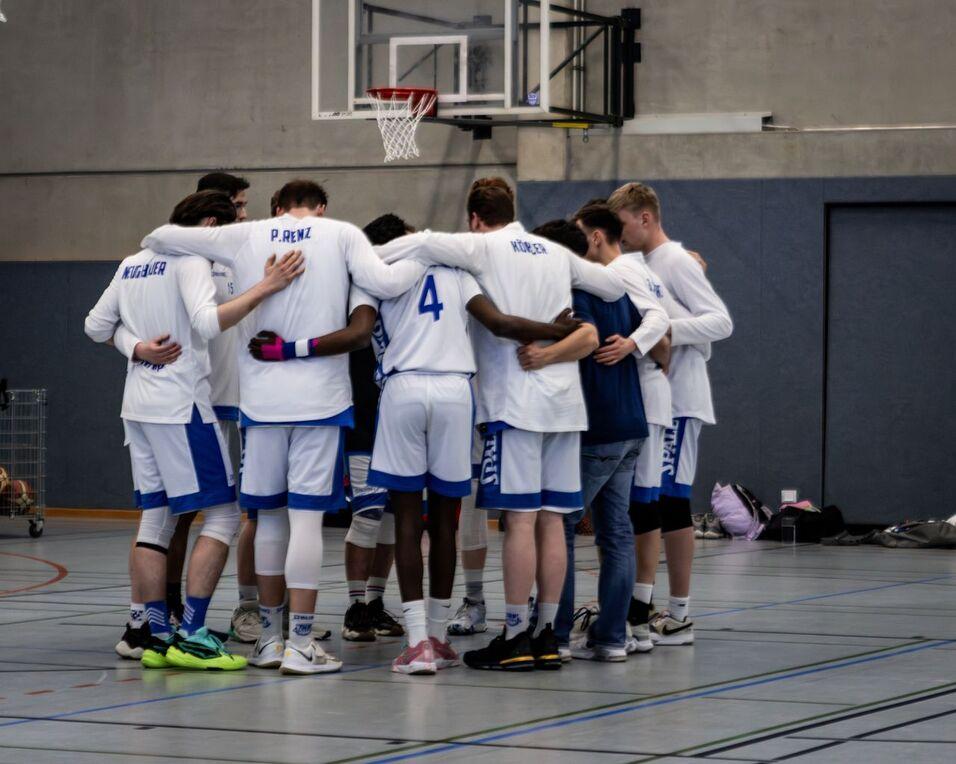 Gruppe von Basketballspielern in weißer und blauer Kleidung umarmt sich in einer Sporthalle vor einem Basketballkorb.