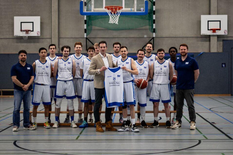 Basketballmannschaft posiert in einer Sporthalle, ein Spieler hält ein Trikot mit der Nummer 20.