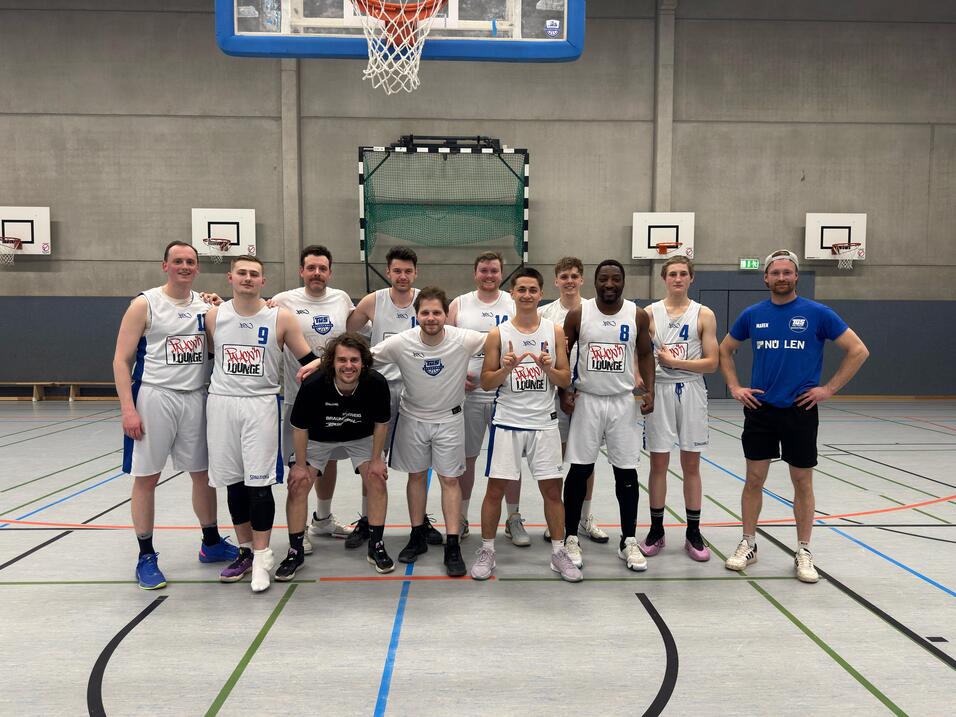 Gruppenfoto von zwölf Basketballspielern in weißen Trikots auf einem Sportplatz, umgeben von Basketballkörben.
