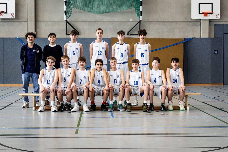 Gruppe von Basketballspielern in weißen Trikots posiert auf einer Bank in einer Sporthalle.