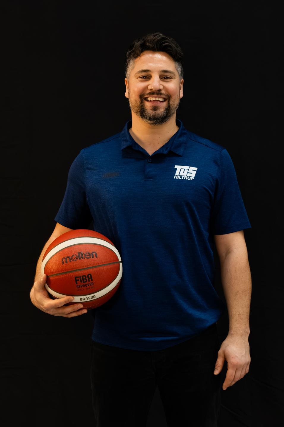 Mann in blauem Polo-Shirt h&auml;lt einen Basketball vor einem schwarzen Hintergrund. L&auml;chelt in die Kamera.