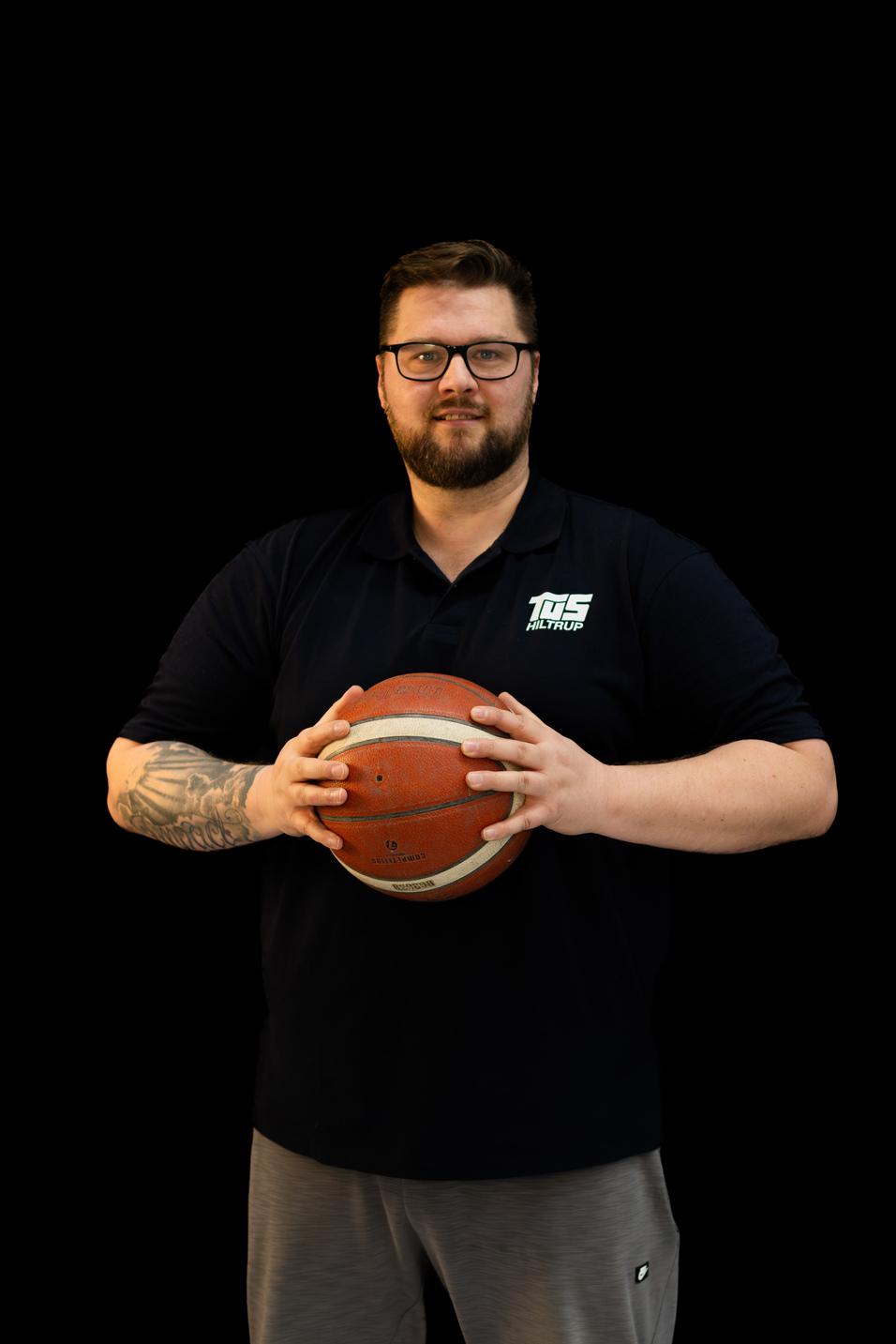 Mann in schwarzer Sportkleidung hält einen Basketball vor einer schwarzen Hintergrundkulisse.