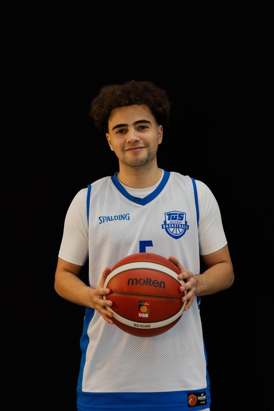 Basketballspieler in Trikot mit Ball vor schwarzem Hintergrund, lächelt in die Kamera.